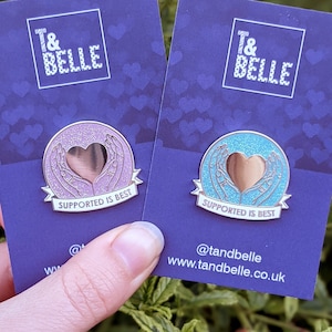 Puede incluir: Dos pines de esmalte con forma de corazón rodeados de manos. Los pines están en tarjetas azules con el texto "T&BELLE" y "SUPPORTED IS BEST". Las tarjetas también incluyen el nombre de usuario de las redes sociales "@tandbelle" y la dirección del sitio web "www.tandbelle.co.uk".