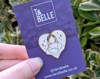 May include: Gold enamel pin featuring a white silhouette of a mother holding a baby inside a heart shape with gold glitter accents. The pin is on a purple background with white hearts. The text "T&BELLE" is at the top of the card and the text "@tandbelle www.tandbelle.co.uk" is at the bottom.