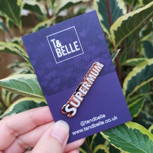 May include: Rose gold enamel pin with the word "Supermum" in white lettering. The pin is on a purple card with white hearts and the text "T&Belle" and "www.tandbelle.co.uk" in white lettering.