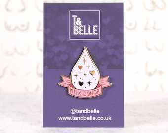 May include: A white enamel pin shaped like a teardrop with gold accents and pink ribbon. The ribbon has the text "Milk Donor" on it. The pin is on a purple card with the text "T&Belle" and the website address "www.tandbelle.co.uk".