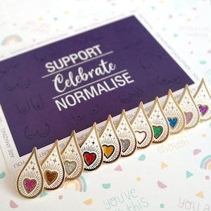 May include: A set of ten gold-coloured enamel pins in the shape of a teardrop with a glitter heart inside. The pins have a white background with a gold outline and a sunburst design. The hearts are different colours: pink, gold, red, orange, silver, green, blue, turquoise, purple, and magenta.