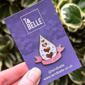 May include: Gold enamel pin featuring a pink ribbon and white teardrop shape with gold hearts and stars. The ribbon has the text "1000 Days" on it. The pin is displayed on a purple background with hearts.