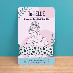 May include: A breastfeeding clothing clip with a black and white polka dot pattern. The product is displayed on a pink and white card with the text "T&BELLE" and "Breastfeeding Clothing Clip". The card features an illustration of a mother breastfeeding her baby.