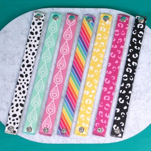 May include: Seven colourful fabric wristbands with different patterns, including polka dots, hearts, rainbows, and animal prints. Each wristband has a silver metal button closure.