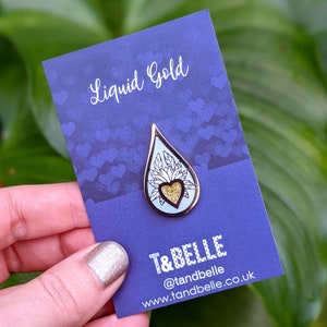 May include: A blue enamel pin with a gold heart and white floral design inside a teardrop shape. The pin says "Liquid Gold" and "T&BELLE" with the website and social media handle below.