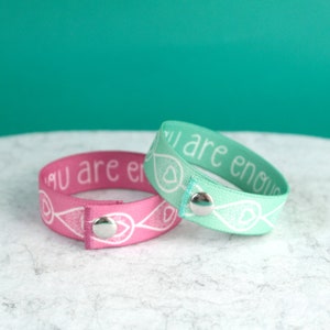 Two breastfeeding clothing clip wristbands with metal clasps. The bracelets are blush pink and mint green and have the text "You are enough" printed in white.