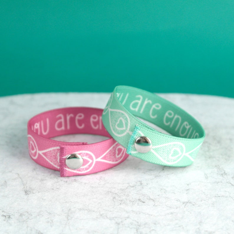 Two breastfeeding clothing clip wristbands with metal clasps. The bracelets are blush pink and mint green and have the text "You are enough" printed in white.