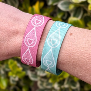 Two breastfeeding clothing clips with a white heart and droplet design, being worn as wristbands. One is pink and the other is mint green.