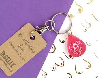 May include: A pink heart-shaped keychain with a white silhouette of a mother breastfeeding a baby. The keychain is attached to a silver keyring and a brown tag that reads "Breastfeeding Love Keepsake Charm" and "T&Belle tandbelle.co.uk".