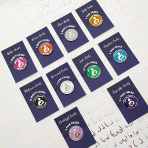 A set of ten glittery 'Breastfeeding Milestone Award' round enamel pin badges. The enamel pins are packaged on purple backing cards showing the milestone for each pin colour from 1 month (pink) up to 5 years+ (purple).