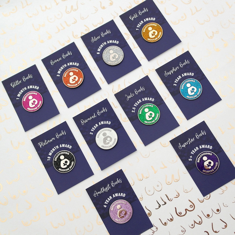 A set of ten glittery 'Breastfeeding Milestone Award' round enamel pin badges. The enamel pins are packaged on purple backing cards showing the milestone for each pin colour from 1 month (pink) up to 5 years+ (purple).