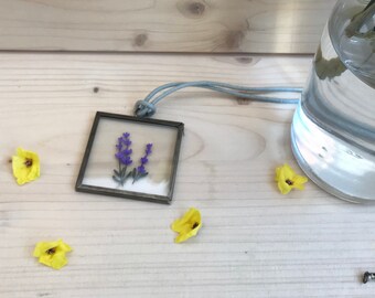 May include: A silver necklace with a square pendant featuring a pressed purple flower inside. The necklace is lying on a wooden surface with yellow flowers scattered around it.