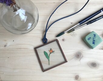 Op de afbeelding: Een ketting met een vierkant frame met een geperste bloem erin. De bloem is een paradijsvogel met oranje, gele en groene bloemblaadjes. Het frame is koperkleurig en de ketting is een donkerblauw koord.