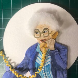 May include: A papercraft illustration of a woman with white hair wearing a blue cardigan over a striped shirt and holding a yellow rotary phone.
