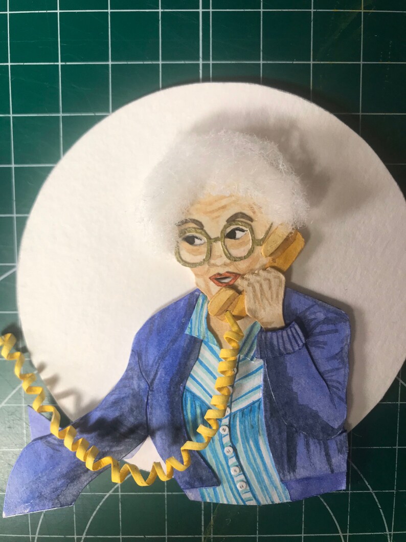 May include: A papercraft illustration of a woman with white hair wearing a blue cardigan over a striped shirt and holding a yellow rotary phone.