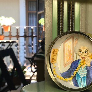 May include: A round, yellow and white tin can with a scene inside. The scene depicts a woman with gray hair talking on a yellow rotary phone. She is wearing a blue and white striped shirt and a blue jacket. The phone cord is yellow and coiled around the woman's arm.