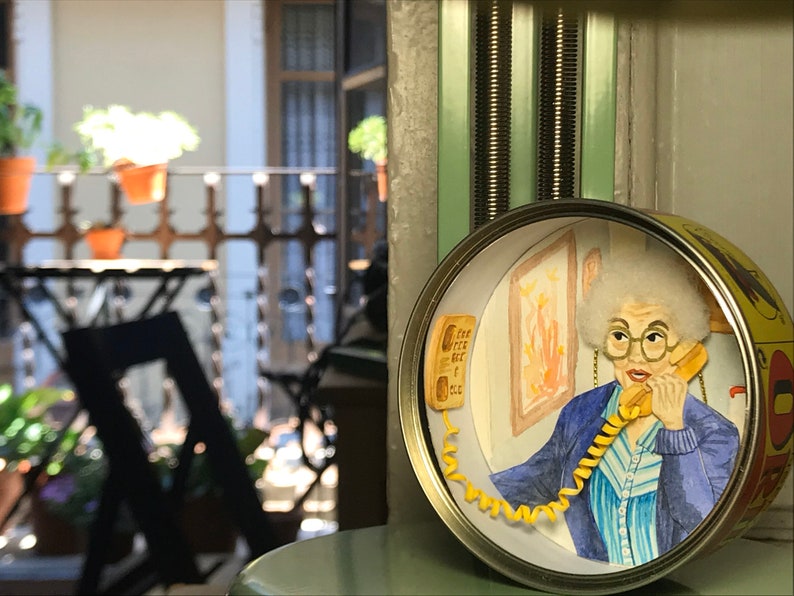 May include: A round, yellow and white tin can with a scene inside. The scene depicts a woman with gray hair talking on a yellow rotary phone. She is wearing a blue and white striped shirt and a blue jacket. The phone cord is yellow and coiled around the woman's arm.
