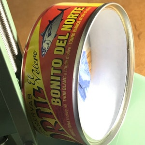 May include: A red, yellow, and white can of Bonito del Norte white tuna in olive oil. The can has a picture of a tuna fish on it. The text on the can reads "Conservas El Velero Bonito del Norte White Tuna in olive oil - Thon Blanc &agrave; l'huile d'olive - Tonno Bianco all'olio di oliva."