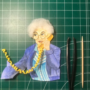 May include: A papercraft figure of a woman with white hair wearing a blue jacket and a striped shirt, talking on a yellow rotary phone with a curly yellow cord.