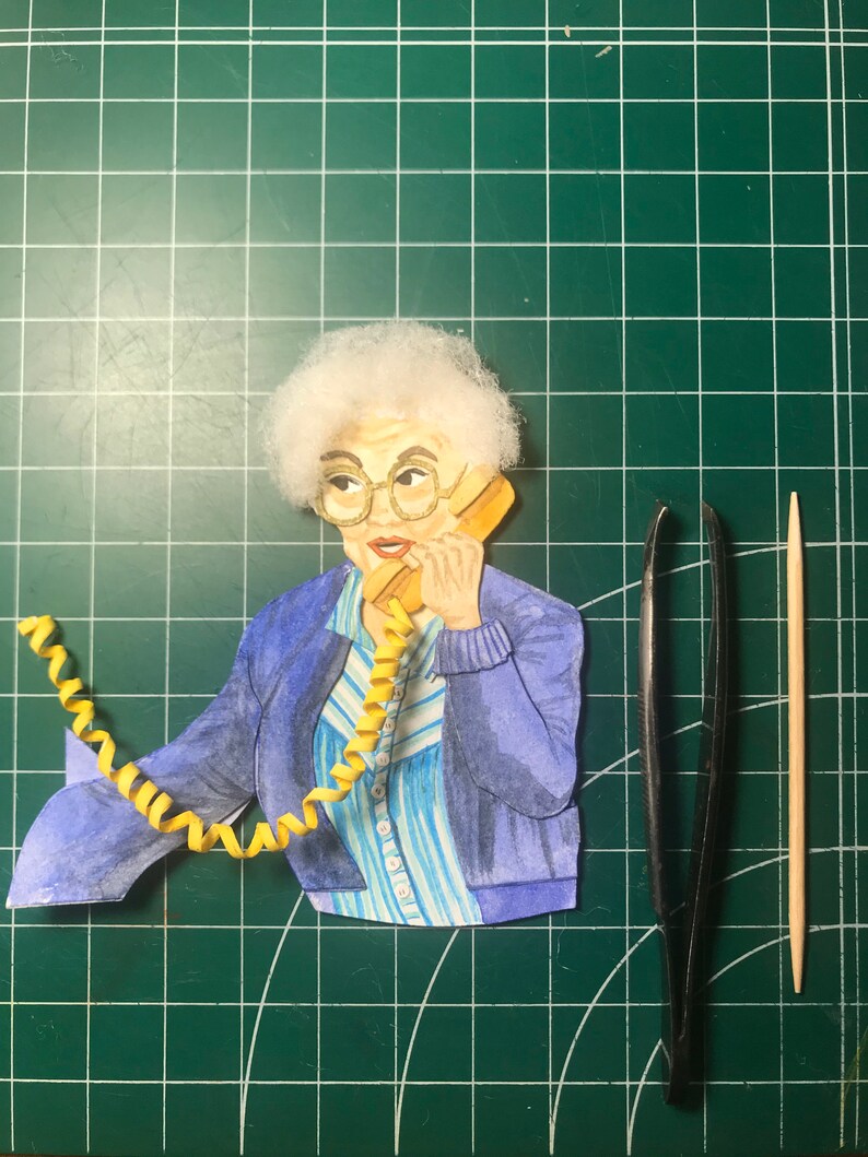 May include: A papercraft figure of a woman with white hair wearing a blue jacket and a striped shirt, talking on a yellow rotary phone with a curly yellow cord.