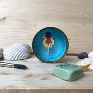 May include: A blue and white tin can with a painted image of a woman in a red swimsuit and a blue hat, holding a yellow ring float. The tin can is sitting on a wooden surface with seashells and a green eraser.