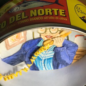 May include: A yellow and red can with the text "O DEL NORTE" and "TONNO BIANCO all'olio di oliva" in a white circle. A woman with glasses is looking at the camera and holding a yellow phone cord. The background is a blue and white striped shirt.