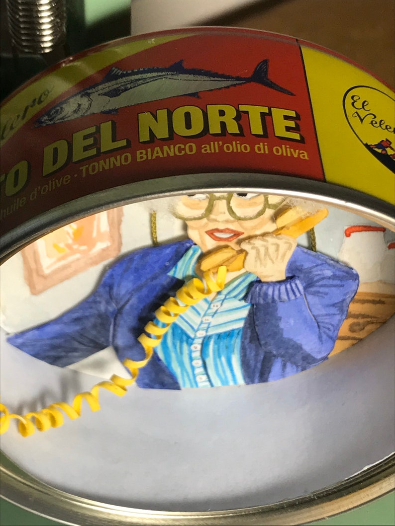 May include: A yellow and red can with the text "O DEL NORTE" and "TONNO BIANCO all'olio di oliva" in a white circle. A woman with glasses is looking at the camera and holding a yellow phone cord. The background is a blue and white striped shirt.