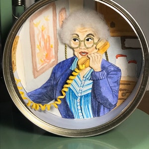 May include: A hand-painted diorama of a woman with white hair wearing a blue sweater and a blue and white striped shirt, talking on a yellow rotary phone. The diorama is inside a metal can.