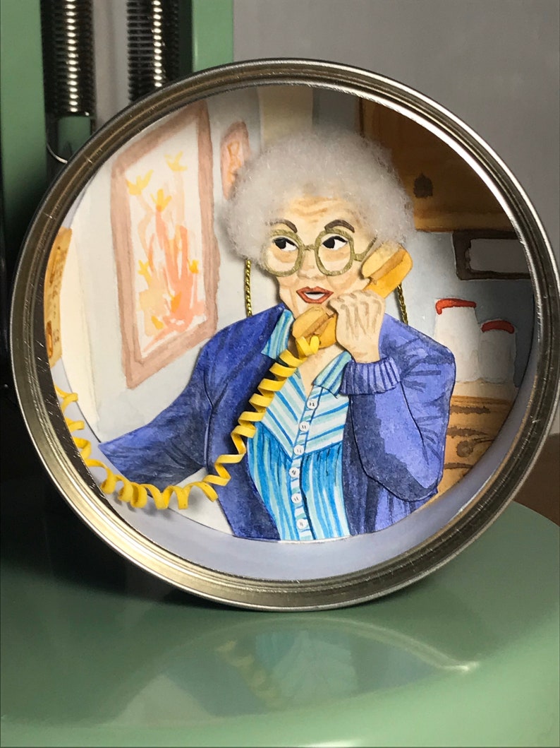 May include: A hand-painted diorama of a woman with white hair wearing a blue sweater and a blue and white striped shirt, talking on a yellow rotary phone. The diorama is inside a metal can.