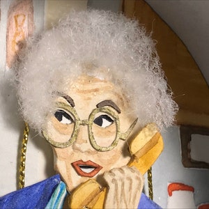 May include: A close-up of a papercraft figurine of a woman with white hair, wearing glasses and a blue jacket, talking on a yellow rotary phone.