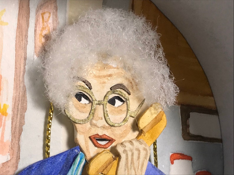 May include: A close-up of a papercraft figurine of a woman with white hair, wearing glasses and a blue jacket, talking on a yellow rotary phone.