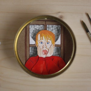 May include: A close-up of a round, metal container with a painted image of a young boy with red hair, wearing a red sweater, looking surprised with his hands on his cheeks. The image is set against a background of a window with white snow outside.
