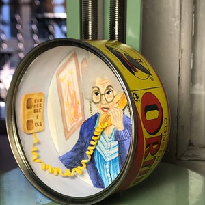 May include: A yellow and red can with a woman's face on it. The woman is wearing a blue sweater and a striped shirt. She is holding a yellow phone with a curly cord. The can says "CONSERVAS ORI" on it.