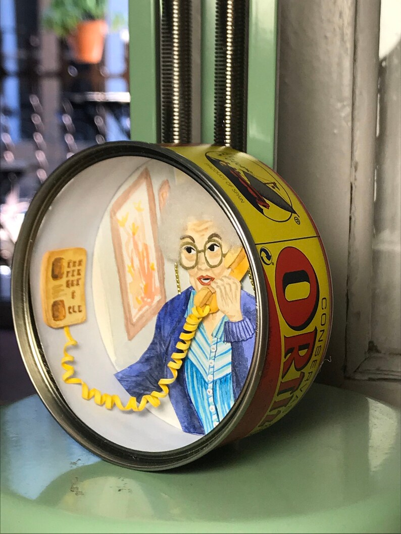 May include: A yellow and red can with a woman's face on it. The woman is wearing a blue sweater and a striped shirt. She is holding a yellow phone with a curly cord. The can says "CONSERVAS ORI" on it.