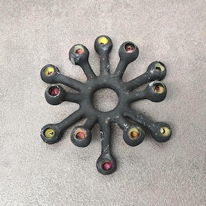 Può includere: Oggetto decorativo a forma di stella grigio scuro, con un foro centrale e bracci radiali. Ogni braccio termina con una forma bulbosa, contenente accenti gialli e rossi. L'oggetto ha una superficie strutturata e sembra essere in metallo.