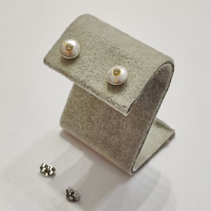 May include: A pair of pearl earrings with a small gold accent on each earring. The earrings are displayed on a gray velvet stand.