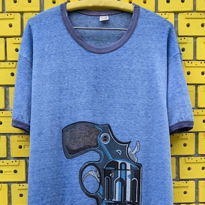 Puede incluir: Una camiseta azul con un gr&aacute;fico de un rev&oacute;lver en la parte delantera. La camiseta tiene un cuello redondo acanalado azul oscuro.