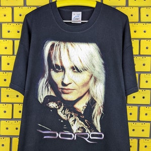 Puede incluir: Camiseta negra con una imagen impresa de una mujer con cabello rubio sosteniendo una serpiente. El texto "DORO" está impreso debajo de la imagen.