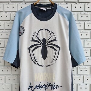 May include: White t-shirt with a blue and light blue colour block design. The shirt features a large black spider graphic with the text "MARVEL by Johnny Blaze" and a small "Spider-Man" logo.