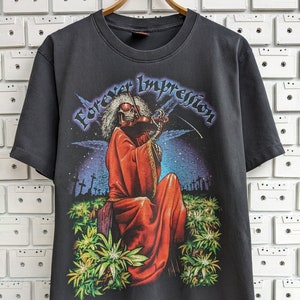 May include: A black t-shirt featuring a graphic print of a skeleton playing a violin in a field of green plants. The text "Forever Impression" is printed in white on the shirt.