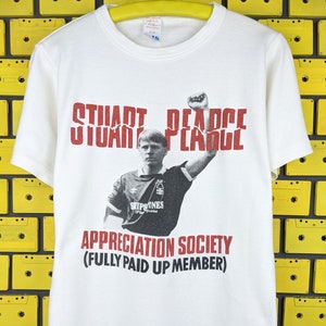 May include: White t-shirt with a red and black graphic of Stuart Pearce, a former English footballer, with the text "Stuart Pearce Appreciation Society (Fully Paid Up Member)".
