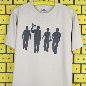 Puede incluir: Una camiseta beige con una silueta negra de cuatro personas. La persona de la izquierda sostiene una maraca.