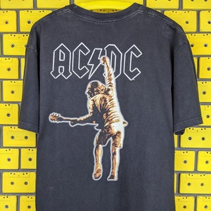 May include: Black t-shirt with a graphic print of a rock band member playing a guitar. The band's name "AC/DC" is printed in white above the graphic.