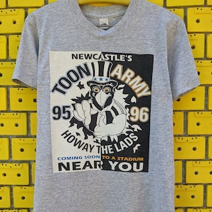 Puede incluir: Camiseta gris con un estampado gr&aacute;fico en blanco y negro de un p&aacute;jaro de dibujos animados con un sombrero con estrellas y rayas. El gr&aacute;fico dice "Newcastle's Toon Army 95-96 Howay the Lads Coming Soon to a Stadium Near You."