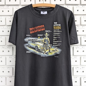 May include: Dark gray t-shirt with a graphic featuring an Australian man, a car, and a dog. The text "ENDANGERED" is above the graphic, and "THE AUSSIE YOBBO" is to the right. The shirt is hanging on a wooden hanger.