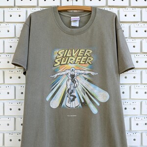 May include: Olive green t-shirt featuring a graphic of the Silver Surfer surfing. The graphic includes the text "THE SILVER SURFER" in yellow and blue, with the character in silver. The shirt is hanging on a wooden hanger.
