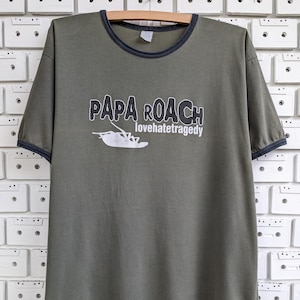 Könnte beinhalten: Olivgrünes T-Shirt mit marineblauen Abschlüssen an Kragen und Ärmeln. Auf der Vorderseite steht der Bandname "PAPA ROACH" in Weiß, darunter "lovehatetragedy" und eine Käfer-Grafik.