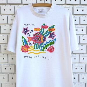 May include: White t-shirt with a colorful "Florida Under the Sea" graphic. The design features a large fish, starfish, and other sea elements in red, yellow, blue, and green. The shirt has a crew neck and short sleeves.