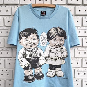 Può includere: T-shirt azzurra con un'illustrazione a fumetti di due figure e la scritta "I LOVE TO ROCK MY GOLD FOR THE LADIES!!". La maglietta ha un girocollo e maniche corte. Il logo Stussy è visibile.