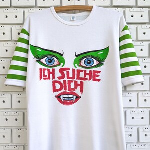 Könnte beinhalten: Weißes T-Shirt mit grün-weiß gestreiften Ärmeln und Besatz. Das Shirt zeigt eine Grafik eines Vampirgesichts mit blauen Augen, grünem Augen-Make-up und dem deutschen Text "ICH SUCHE DICH" in Rot.